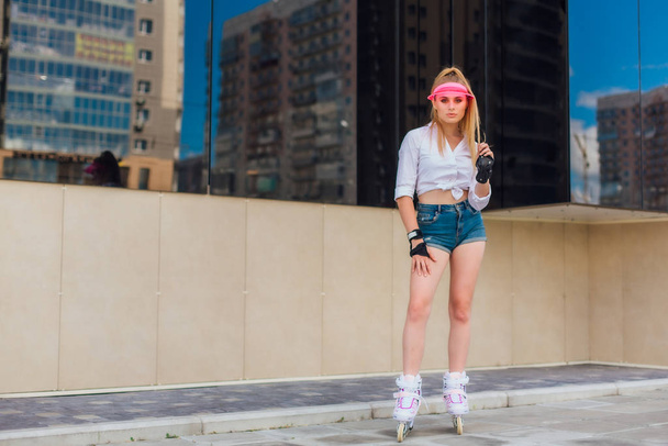 Portrait of an emotional girl in a pink cap visor and protective gloves for rollerblades and skateboarding riding on rollerblades on the road. - Photo, image