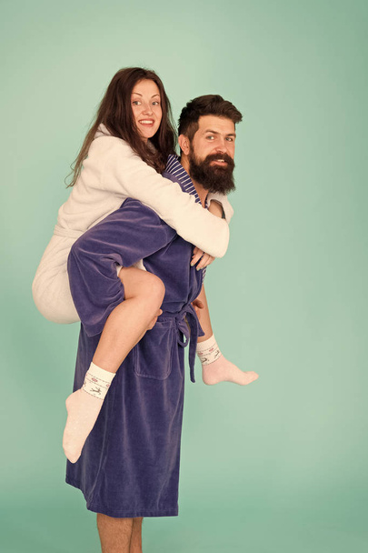 Handsome young man piggybacking beautiful woman. Couple in bathrobes having fun turquoise background. Lets stay at home and have fun. They always have fun together. Enjoying every second together - Zdjęcie, obraz