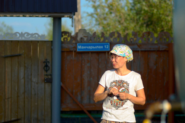Посміхаючись Якутська дівчина в футболці і шапці знаходиться на житловій ділянці приватного будинку в північному селі Росії. - Фото, зображення