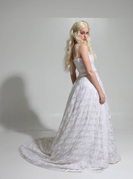 full length portrait of blonde girl wearing long white lace bridal gown. standing pose on a studio background. - Photo, Image