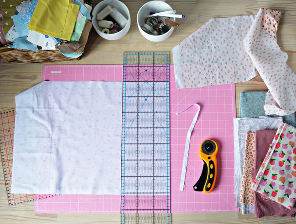 Work place: cotton fabrics, pink cutting mat, quilting ruler, rottary cutter, sewing supplies in white cups and wooden desk - Fotoğraf, Görsel