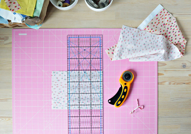 Work place: cotton fabrics, pink cutting mat, quilting ruler, rottary cutter, sewing supplies in white cups and wooden desk - Foto, immagini