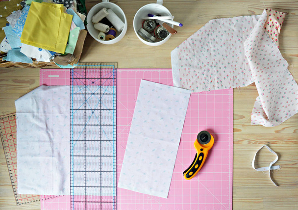 Work place: cotton fabrics, pink cutting mat, quilting ruler, rottary cutter, sewing supplies in white cups and wooden desk - Foto, imagen