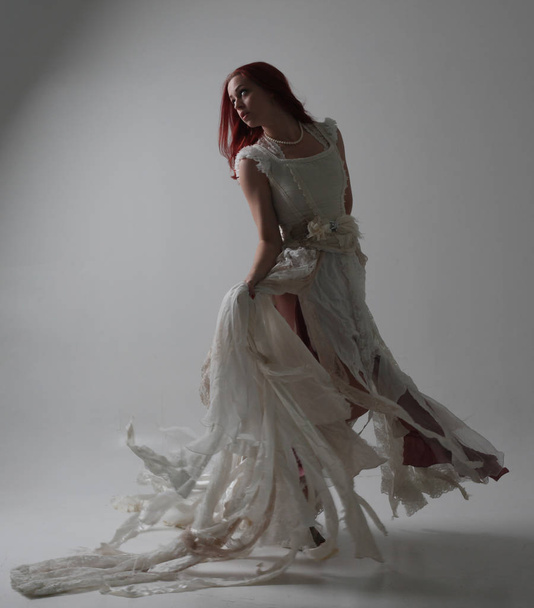 full length portrait of red haired girl wearing torn and tattered wedding dress. Standing pose against a studio background with contrasty shadow lighting. - Photo, Image