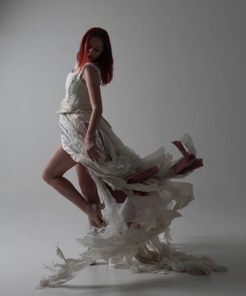 full length portrait of red haired girl wearing torn and tattered wedding dress. Standing pose against a studio background with contrasty shadow lighting. - Photo, image