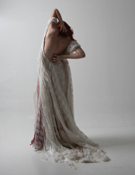 full length portrait of red haired girl wearing torn and tattered wedding dress. Standing pose against a studio background with contrasty shadow lighting. - Fotó, kép