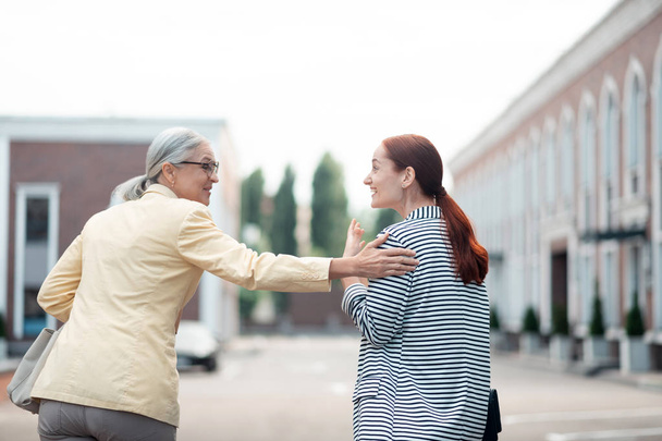 Friendly gesture. Senior Caucasian businesswoman standing outdoors while laughing and patting her young female colleague on the shoulder - Photo, image