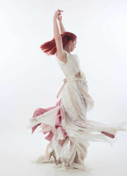 full length portrait of red haired girl wearing torn and tattered wedding dress. Standing pose against a  white studio background. - Foto, immagini