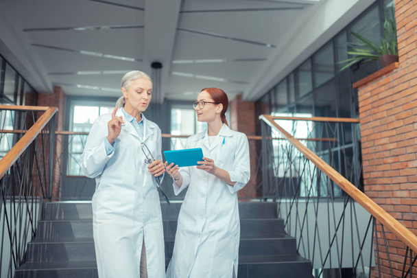 Female doctors. Senior Caucasian female doctor standing on the stairs in a modern medical establishment while talking to her young colleague - Fotoğraf, Görsel