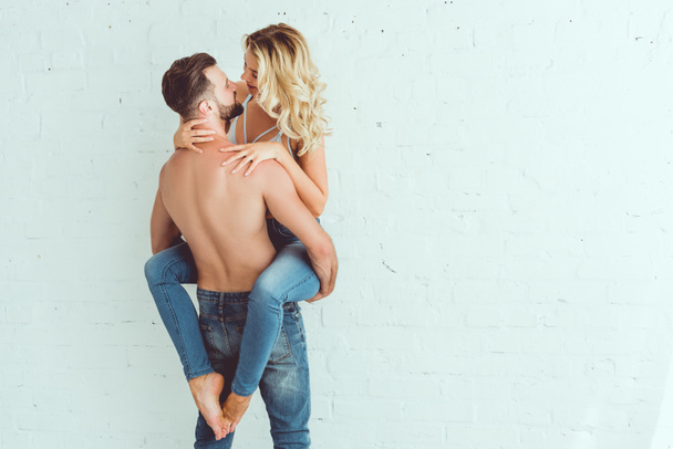 back view of young shirtless man holding on hands beautiful girlfriend while standing near white wall - Foto, imagen