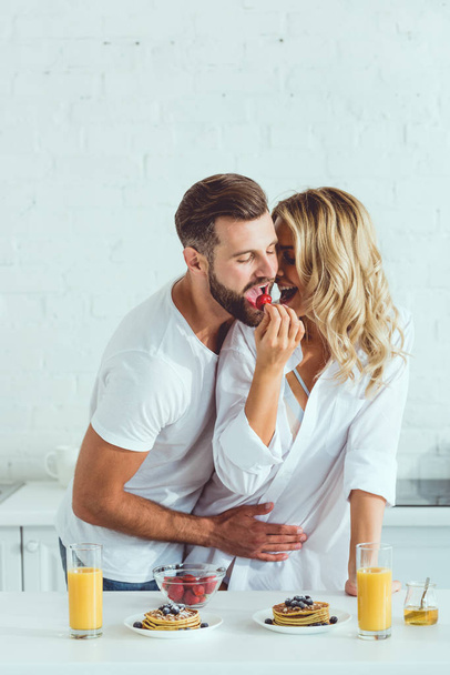 happy young woman feeding boyfriend with strawberry while standing at kitchen table with served breakfast - Photo, Image