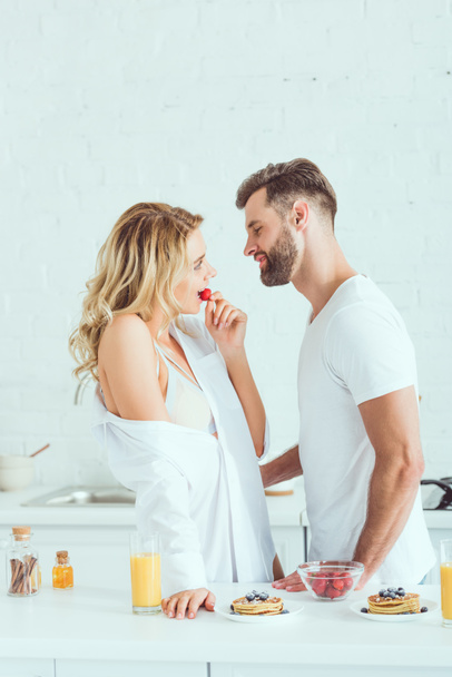happy young man standing near beautiful girlfriend eating strawberry in kitchen - Foto, Imagem