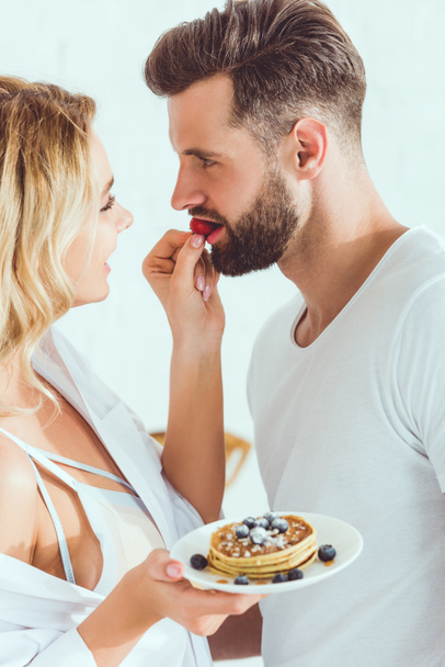 jovem sensual mulher alimentando namorado com morango enquanto segurando prato com panquecas
 - Foto, Imagem