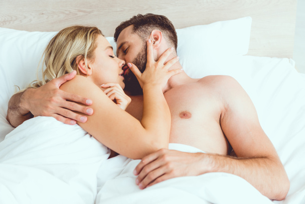 happy young couple hugging and kissing with closed eyes while lying in bed - Zdjęcie, obraz