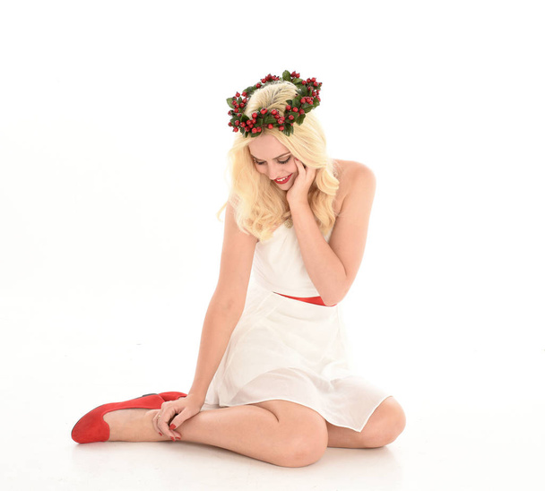 Ganzkörperporträt eines blonden Mädchens, das ein weißes Kleid und eine Blumenkrone trägt. sitzend, isoliert vor weißem Studiohintergrund. - Foto, Bild