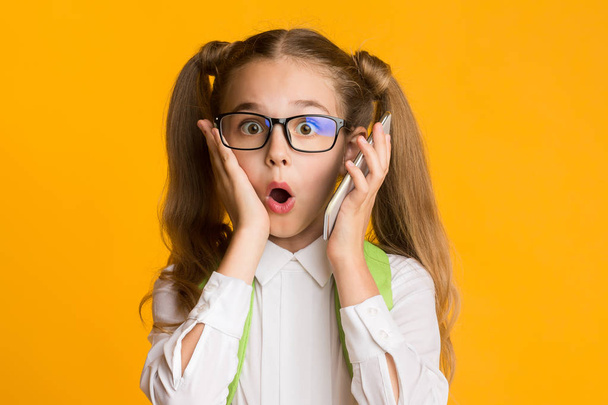 Surprised School Girl Holding Cellphone To Ear On Yellow Background - 写真・画像