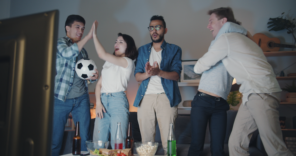テレビでサッカーを見ている陽気なスポーツファンは、自宅で夜にハイファイブを行います - 映像、動画