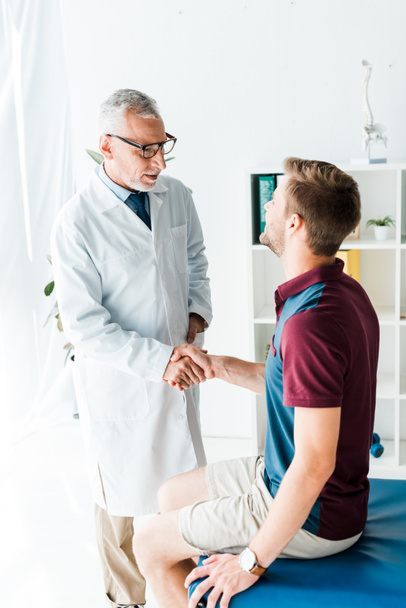 happy doctor in glasses shaking hands with young man in clinic  - 写真・画像