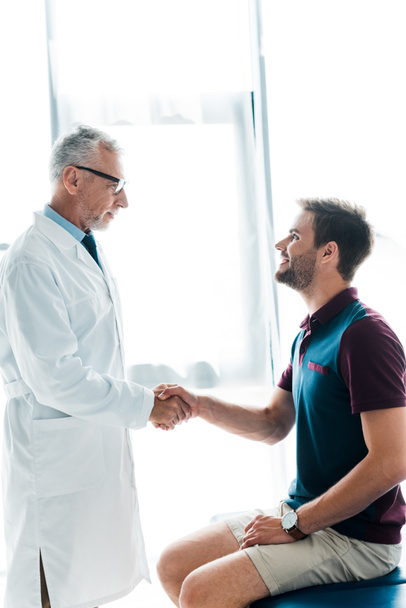 szczęśliwy lekarz w okularach drżenie rąk z wesoły młody człowiek w klinice  - Zdjęcie, obraz