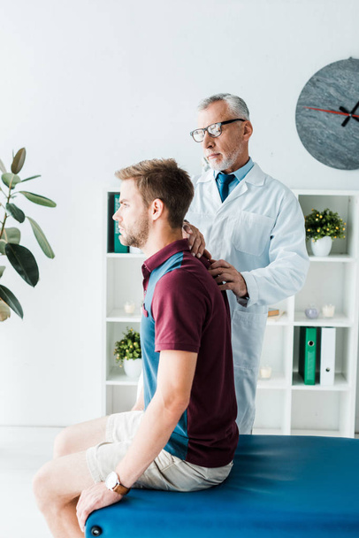 chiropractor in white coat and glasses touching back of handsome patient in clinic  - Fotó, kép
