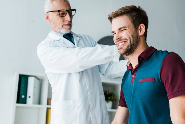 chiropractor in white coat and glasses touching happy patient in clinic  - Foto, afbeelding