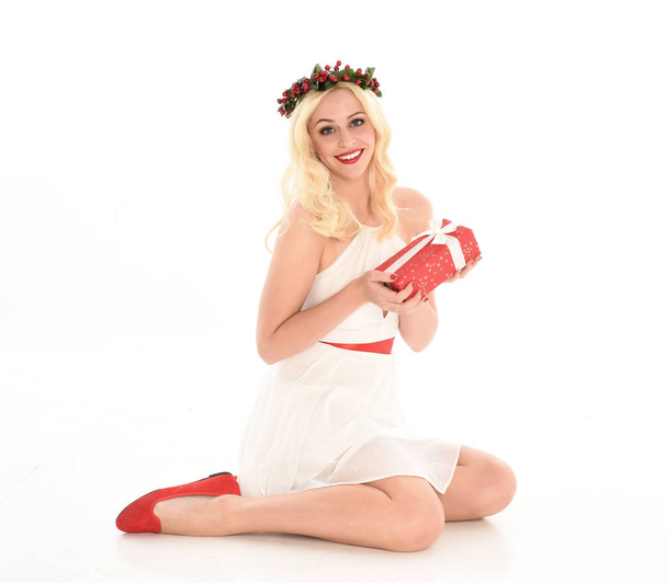 pełnej długości portret blond dziewczyna ubrana w białą sukienkę i koronę kwiatową. Siedząc poza, trzymając czerwony prezent świąteczny, odizolowany od białego tła studyjnej. - Zdjęcie, obraz