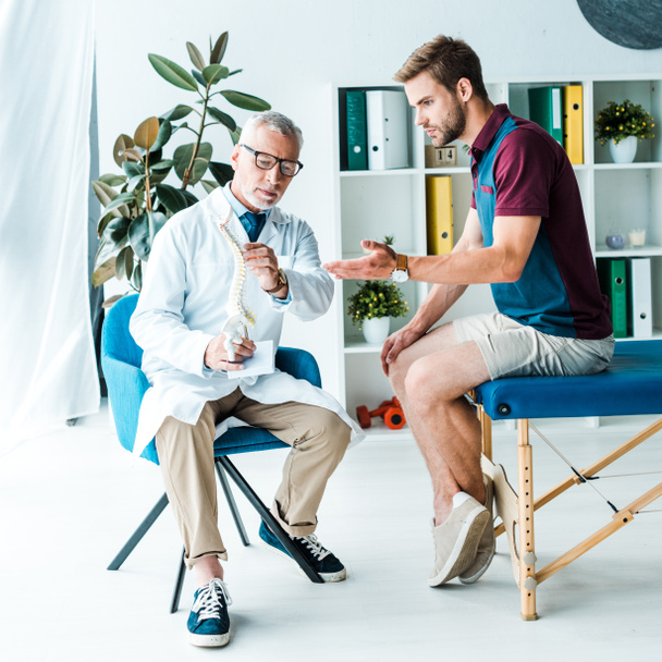 schöner bärtiger Mann gestikuliert beim Anblick eines Wirbelsäulenmodells in der Nähe eines Arztes mit Brille  - Foto, Bild
