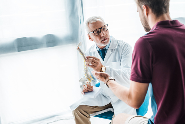 selective focus of bearded doctor in glasses holding spine model near patient  - Φωτογραφία, εικόνα