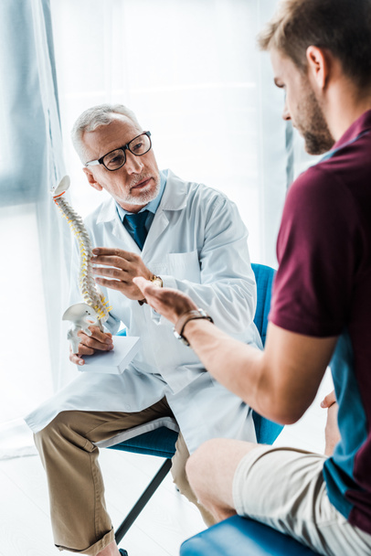 selective focus of bearded doctor in glasses holding spine model near patient gesturing in clinic  - 写真・画像