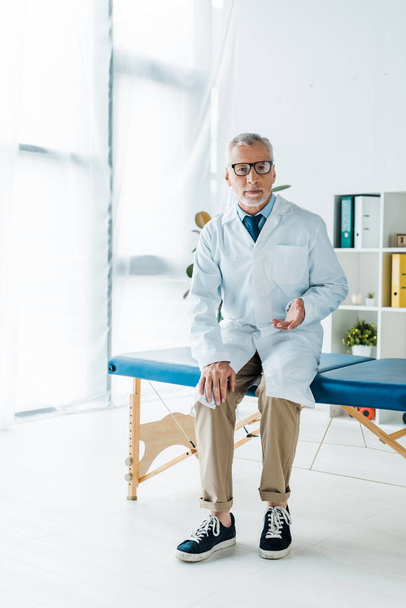 médico barbudo em óculos e casaco branco sentado na mesa de massagem e gesticulando na clínica
  - Foto, Imagem