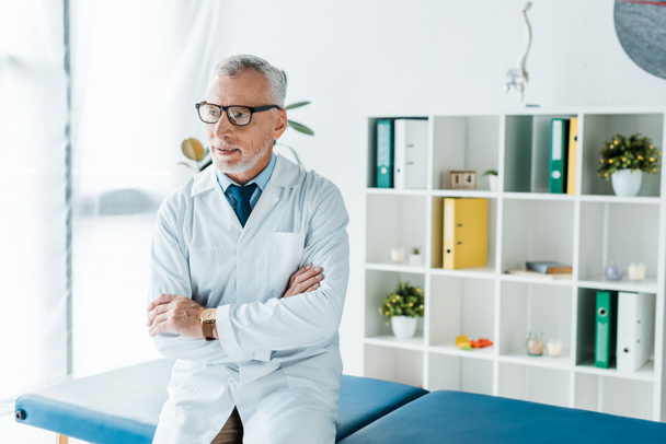 Brodaty lekarz w okularach i biały płaszcz siedzi na stole do masażu z skrzyżowanymi ramionami  - Zdjęcie, obraz