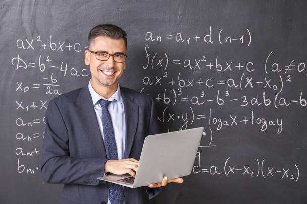 Красивый учитель математики с ноутбуком рядом с доской в классе
 - Фото, изображение