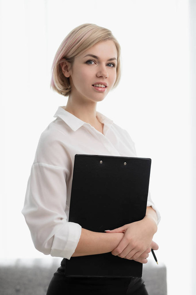 Young beautiful blonde girl with short hair in a white shirt is standing in the office and holding a notebook and pen. - Foto, immagini