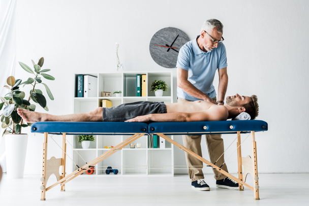  bearded chiropractor doing massage to man on massage table near clock - Photo, Image