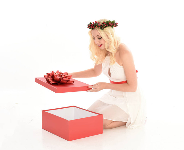 full length portrait of blonde girl wearing a white dress and flower crown.  Sitting pose, holding a red Christmas present, isolated against a  white studio background. - Photo, Image
