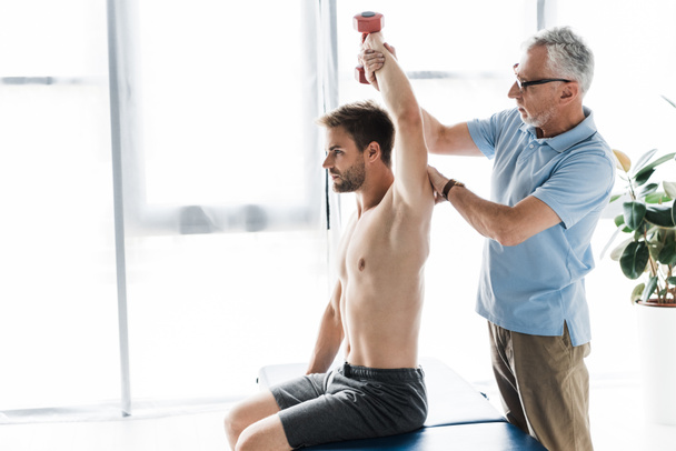 chiropractor standing near man exercising with dumbbell in clinic  - Photo, Image