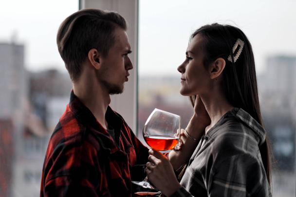 Young beautiful couple in plaid shirts on the balcony of a multistory building. Drink and taste wine, laugh and embrace. - Photo, Image