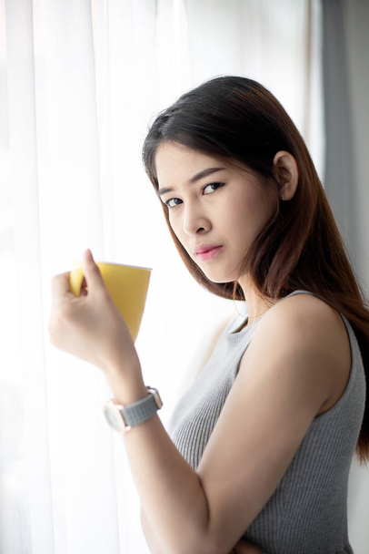 Asian woman holding yellow mug near window with natural light. - Φωτογραφία, εικόνα