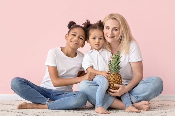 Gelukkige vrouw en haar kleine Afro-Amerikaanse dochters met ananas zittend in de buurt van kleur muur - Foto, afbeelding