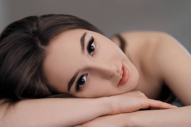 Beautiful brunette girl with brown eyes with bare shoulders lying on the bed. Face close up. Asian and Oriental type of appearance. - Fotografie, Obrázek
