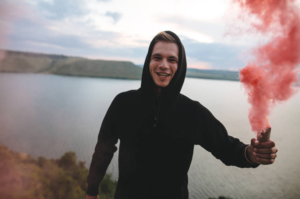 Stylish hipster holding red smoke bomb in hand, standing on top  - Photo, Image