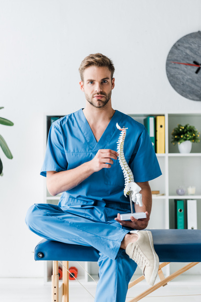 handsome doctor looking at camera while holding spine model in clinic  - Foto, Imagen
