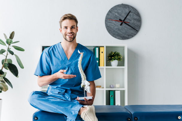 cheerful doctor looking at camera while gesturing and holding spine model in clinic  - Foto, Bild