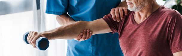 panoramic shot of doctor standing near mature man exercising with dumbbell in clinic  - Photo, Image