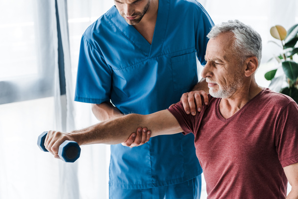 cropped view of doctor standing near mature man holding dumbbell in clinic  - Photo, Image