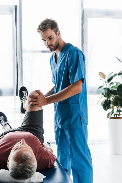 beau médecin touchant jambe de patient mature faisant de l'exercice sur table de massage
  - Photo, image