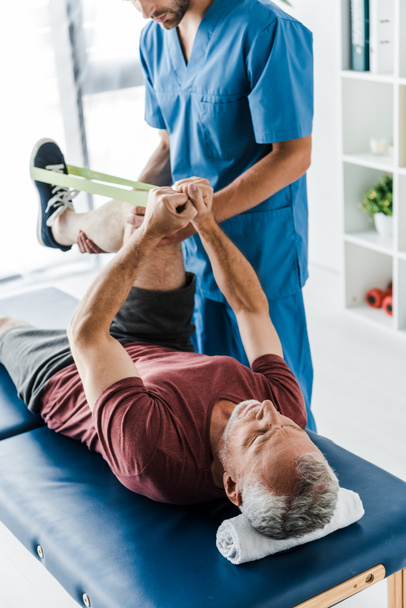 bijgesneden beeld van bebaarde Doctor Holding been van volwassen patiënt trainen met elastiek  - Foto, afbeelding