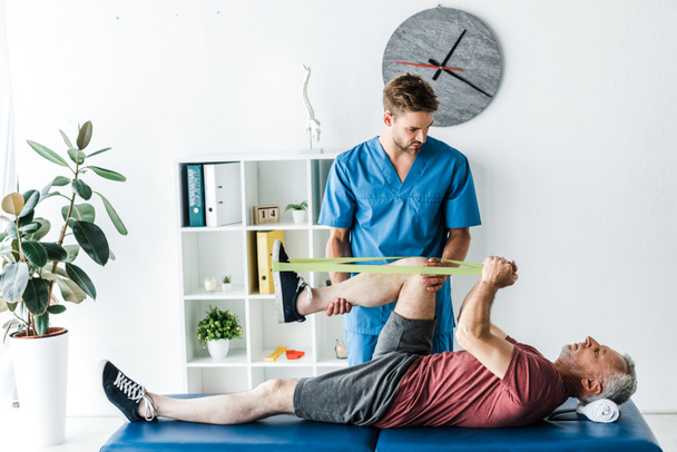 handsome doctor holding leg of mature patient exercising in clinic  - Photo, Image