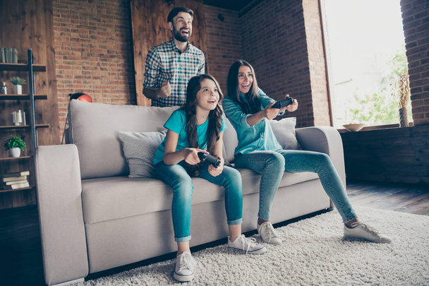 Portrait of excited people and little girl with brunet hair play videogame wearing denim jeans t-shirt sit divan in house indoors - Foto, Imagen