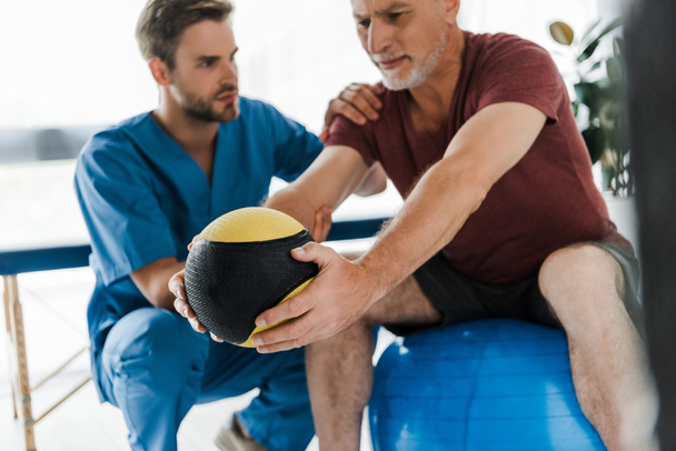 επιλεκτική εστίαση του ώριμου ασθενούς άσκηση σε μπάλα γυμναστικής κοντά στο γιατρό  - Φωτογραφία, εικόνα